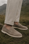 A person wearing light beige trousers and taupe suede slip-on loafers standing outdoors on grassy ground with mountains in the background.
