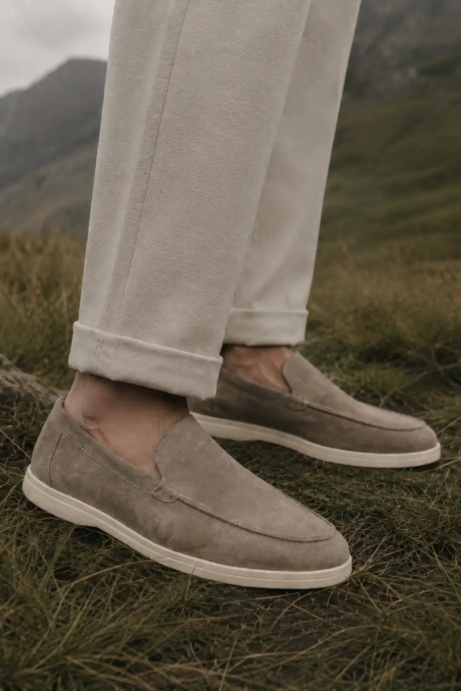 A person wearing light beige trousers and taupe suede slip-on loafers standing outdoors on grassy ground with mountains in the background.