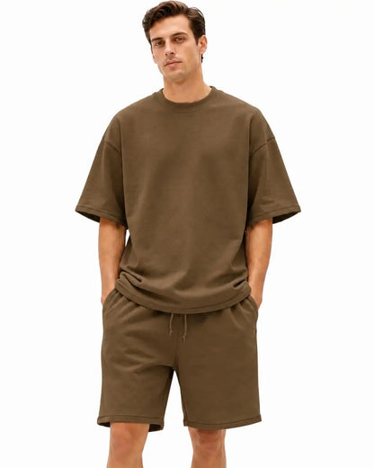 A man modeling a matching two-piece lounge set consisting of an oversized short-sleeve crewneck t-shirt and drawstring shorts in a solid olive brown color, set against a plain white background.