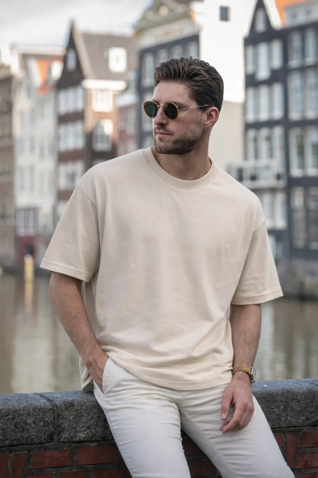 Man wearing a beige t-shirt and sunglasses sitting by a canal with buildings in the background
