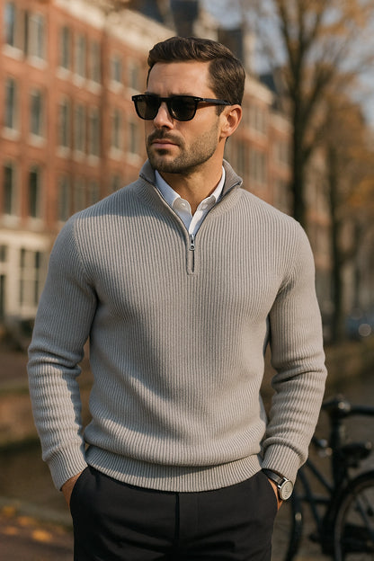 Man wearing a light gray sweater and sunglasses outdoors with buildings in the background