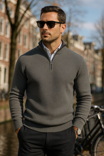 Man wearing a gray sweater and sunglasses standing outdoors with buildings and a bicycle in the background.