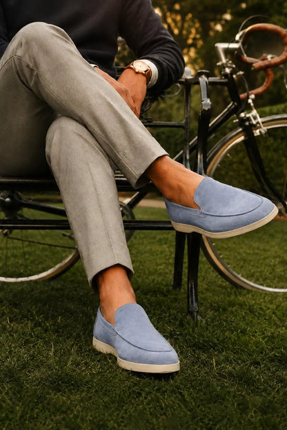 A person sitting on a black bicycle outdoors on grass, wearing light blue suede slip-on loafers with white soles. The person is also wearing light gray trousers and a dark long-sleeve top.