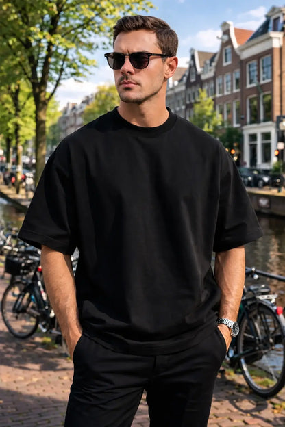 Man wearing a black t-shirt and sunglasses standing in an urban setting with buildings and bicycles.