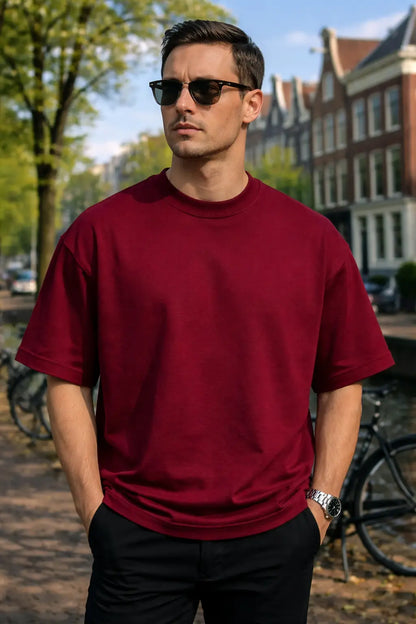 Man wearing a red t-shirt and sunglasses standing outdoors with buildings and trees in the background